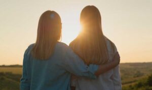 mom with arm around teen daughter silohouetted with rising sun in background