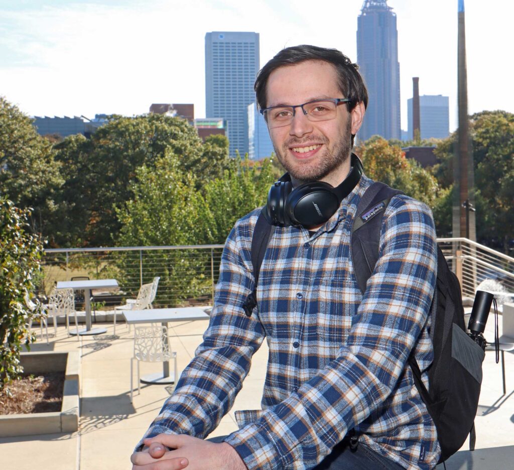 mail college student in plaid flannel shirt wearing backpack seated outdoors with Atlanta skyline in background