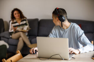 teen wearing headphones with a laptop and adult in background providing support