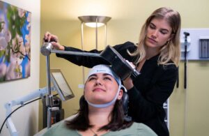 young woman receives TMS therapy, sitting in chair in clinic, wearing cloth cap on head, TMS technician adjusts machine to rest lightly on head