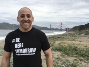 man stand smiling in shirt that reads "be here tomorrow" in front of san francisco bridge