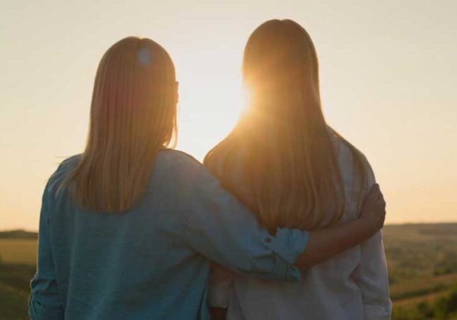 mom with arm around teen daughter silohouetted with rising sun in background